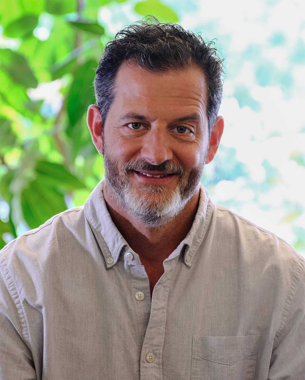 A man with dark hair and a gray beard smiles against lush greenery.